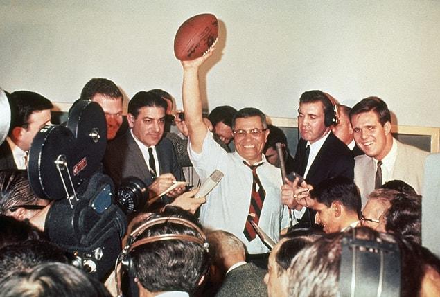 21. Green Bay Packers coach Vince Lombardi raises a football in victory, surrounded by reporters covering the first Super Bowl in 1967.