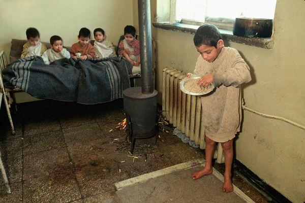 3. A hungry boy stands alone and eats with his hands as other boys sit together under a blanket on a bed beside a small wood-burning stove in a hospital for mentally handicapped children in Kavaja, Albania in March 1992.