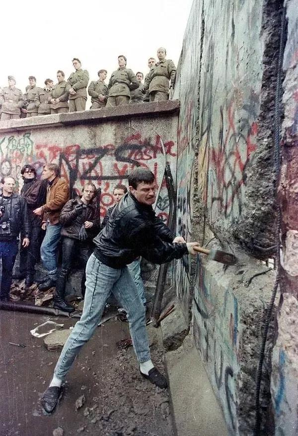 33. Bir gösterici Berlin Duvarı'na balyozla vururken, Doğu Belin sınırındaki görevliler izliyor.