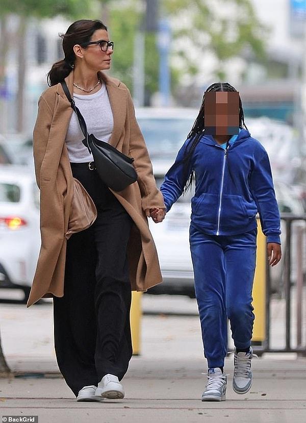 Sandra Bullock beyaz bir üst, bir palto ve siyah bir pantolon, spor ayakkabı giydi ve siyah bir el çantası taktı.