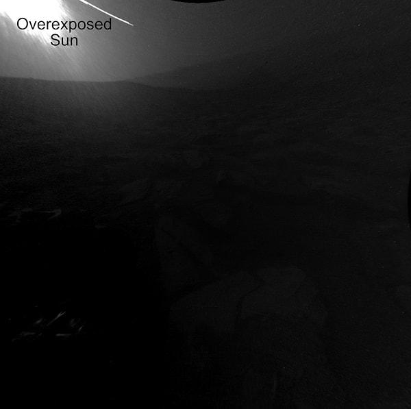 Gündoğumu sırasında gökyüzü aydınlandıkça, izcinin 2 metre uzunluğundaki kolunun gölgesi sola doğru hareket ediyor ve Curiosity'nin ön tekerlekleri karenin her iki tarafındaki karanlıktan görülüyor.