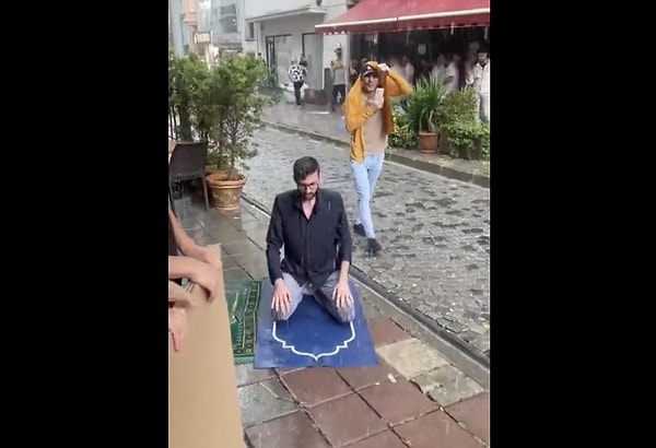 Bir vatandaş yağmur altında namaz kılarken iki kişi de ellerinde kartonla namaz kılan kişinin ıslanmasını engellemeye çalıştı.